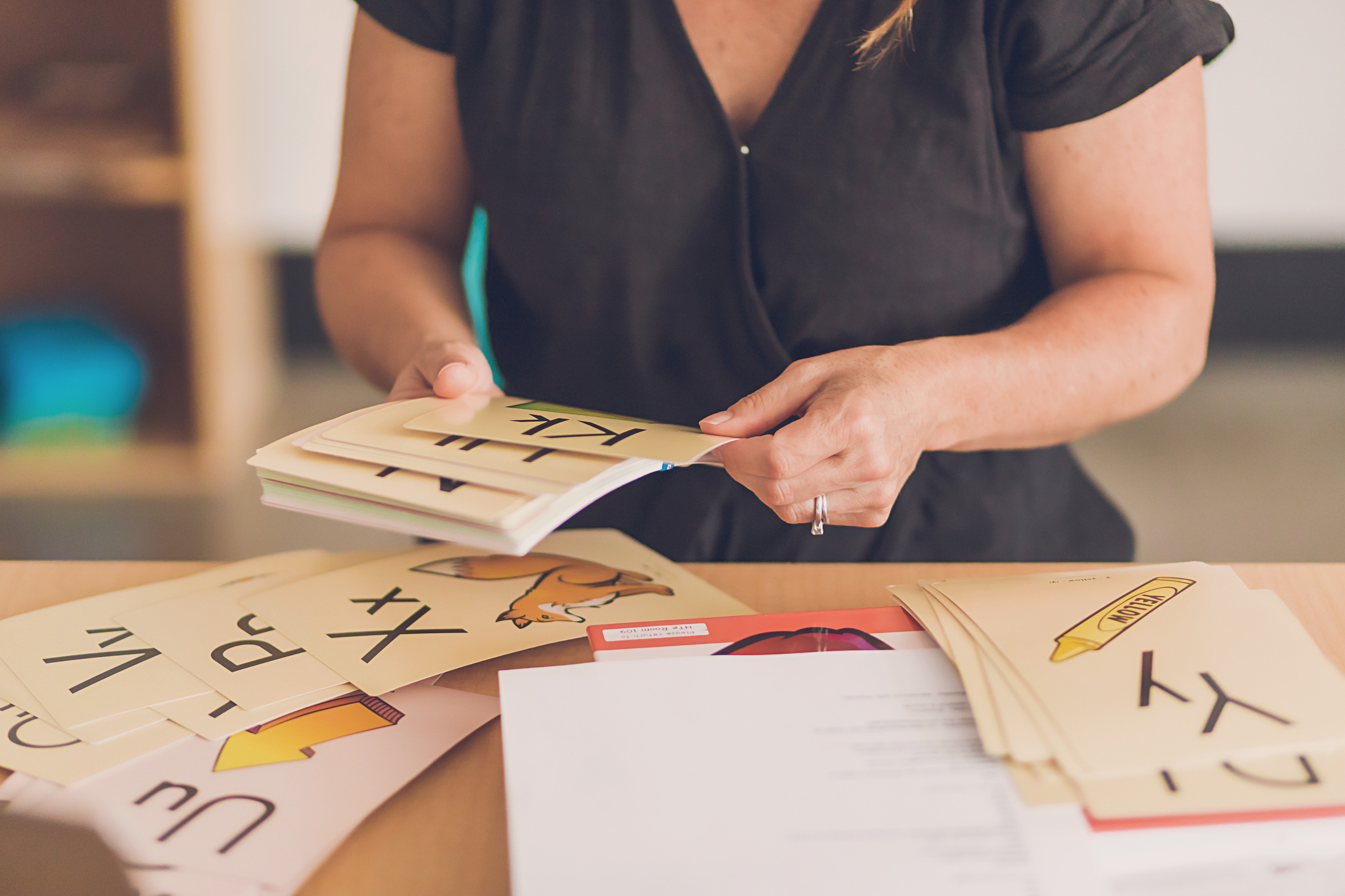 Professora organizando cartões educativos com letras e ilustrações sobre uma mesa durante atividade de alfabetização e letramento infantil.