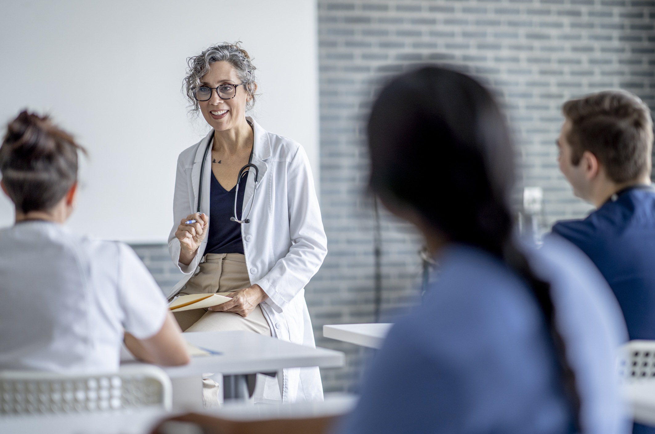 Médica de jaleco branco e estetoscópio, sorrindo enquanto explica algo para um grupo de estudantes sentados em sala de aula, em um ambiente moderno e iluminado.