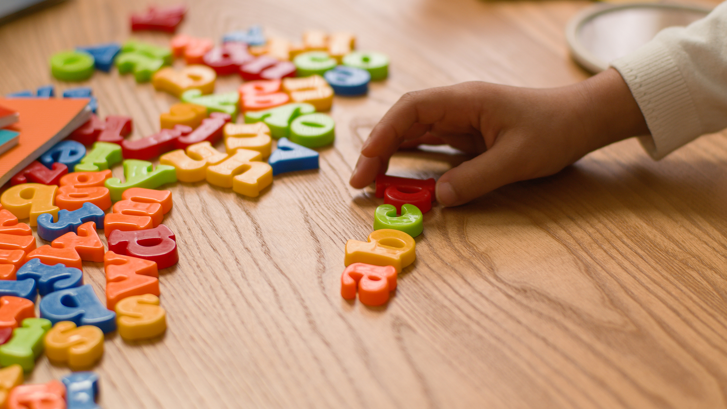 Mão de criança organizando letras coloridas sobre mesa de madeira durante atividade de alfabetização infantil.