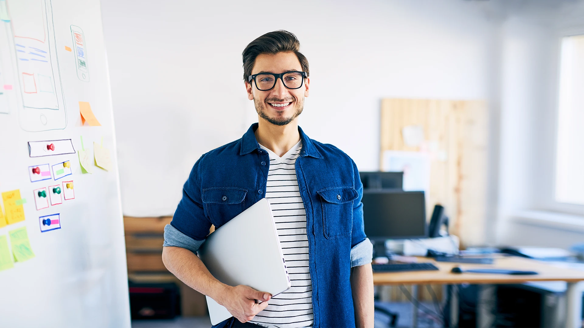 Jovem designer ou desenvolvedor sorridente segurando um notebook em um escritório moderno com um quadro branco ao fundo