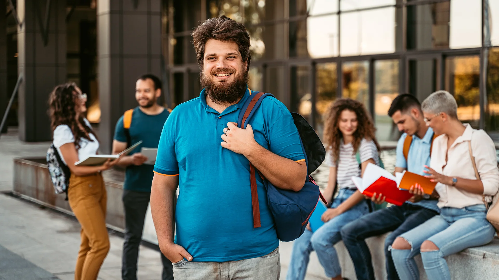 Estudante universitário sorridente com mochila em frente ao campus da faculdade com colegas ao fundo