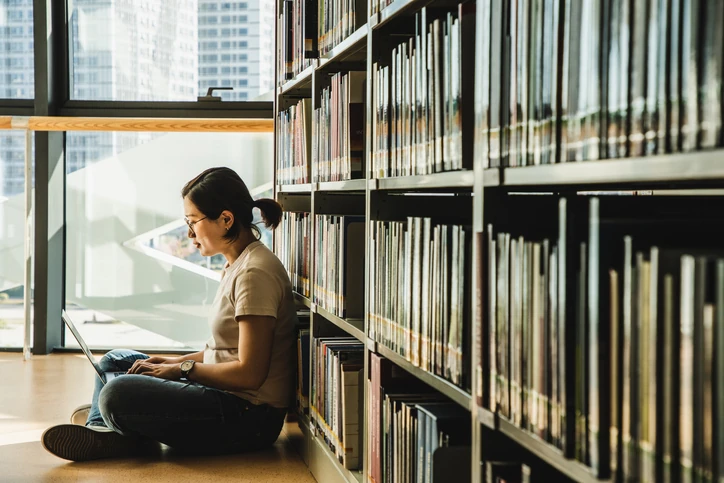 Estudante focada realizando pesquisa acadêmica em notebook dentro de uma biblioteca moderna com estantes de livros