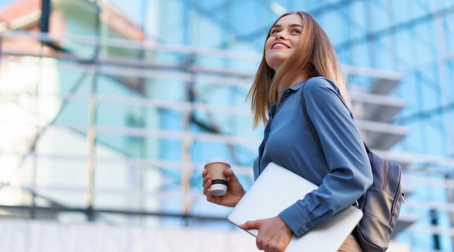 Estudante universitária sorridente caminhando no campus com notebook e café