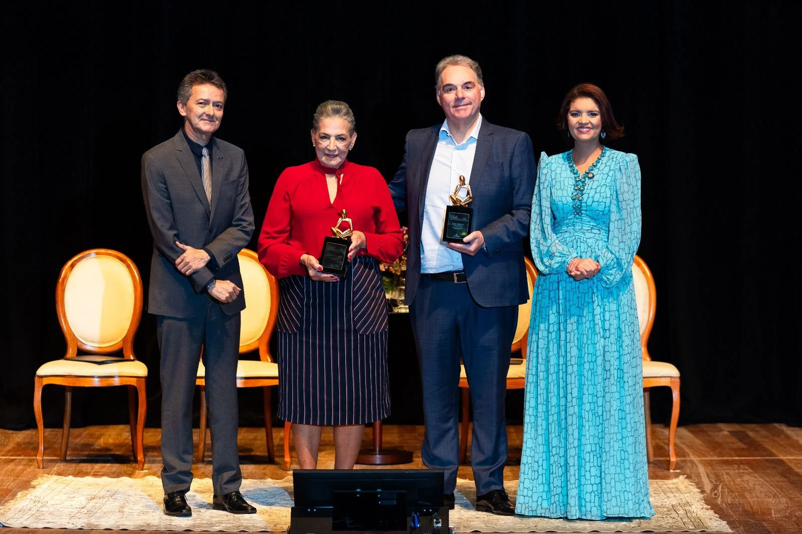 Quatro pessoas em pé no palco recebendo troféus de premiação em um evento formal.