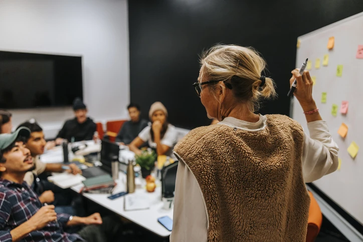 Professora experiente utilizando lousa branca com notas adesivas em aula colaborativa com estudantes universitários