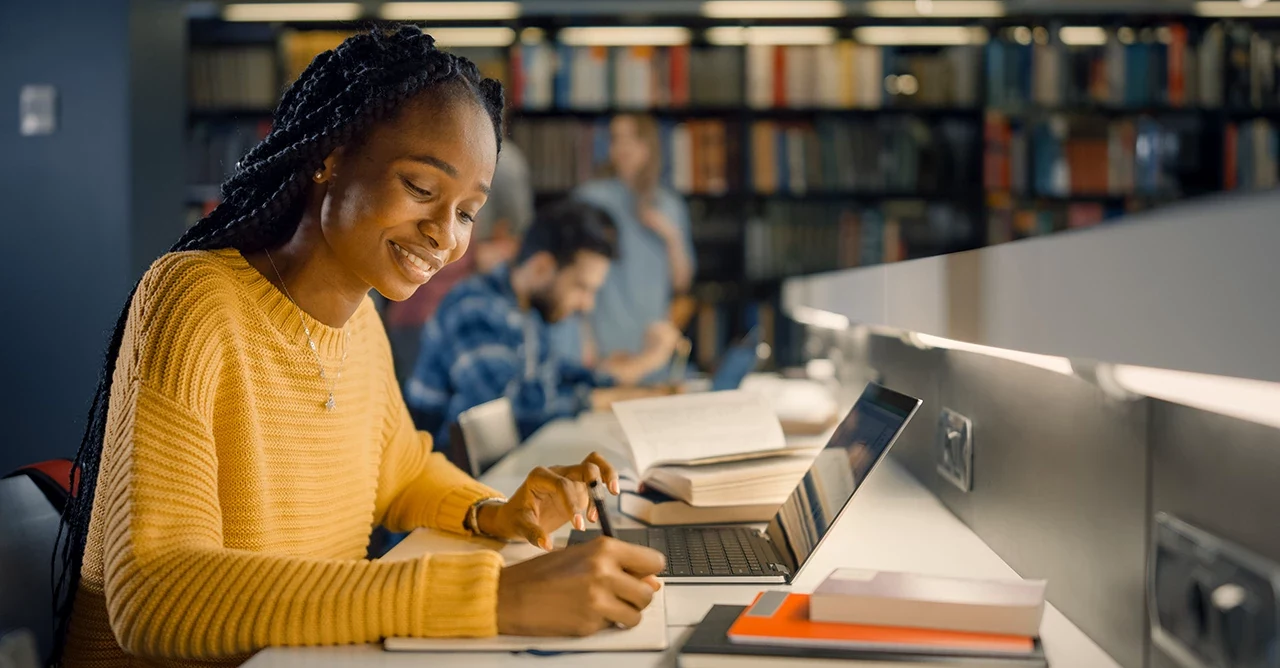 Estudante negra sorridente escrevendo em um caderno em uma biblioteca universitária com laptop