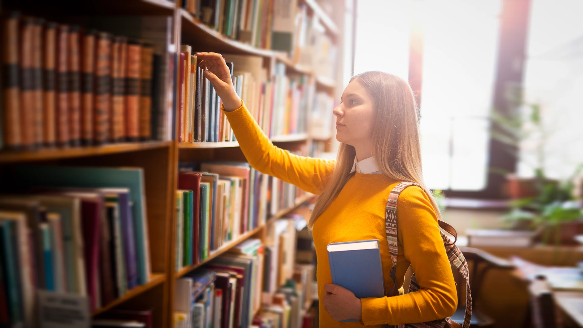 Estudante loira escolhendo um livro em uma prateleira de biblioteca universitária