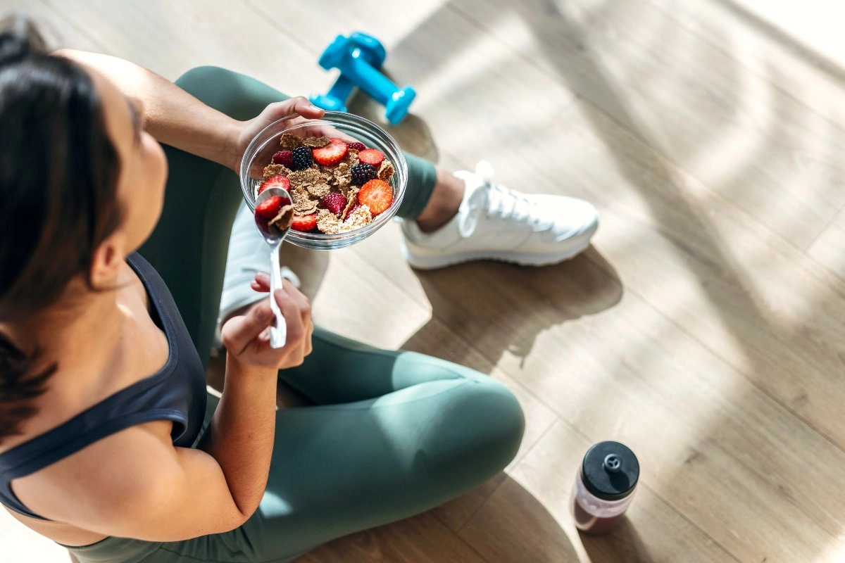 Mulher vestindo roupas esportivas comendo uma tigela de cereais com frutas após o treino Importância da nutrição esportiva para a recuperação muscular e melhor desempenho físico nos treinos