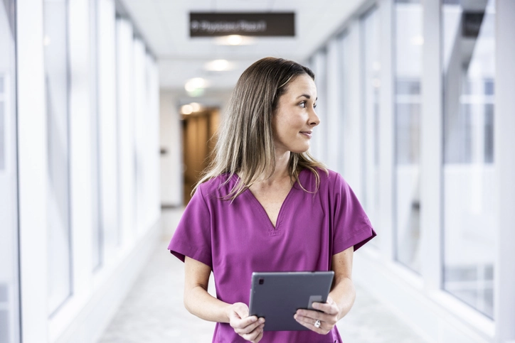 Descubra quanto tempo dura a faculdade de enfermagem e conheça a rotina profissional na área da saúde Estudante ou profissional de enfermagem vestindo uniforme roxo segurando um tablet em um corredor de hospital