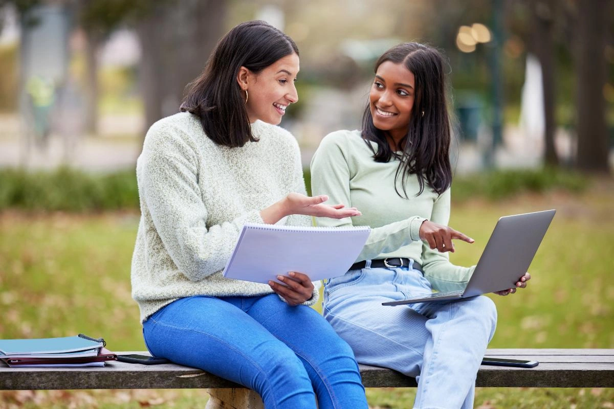 Duas estudantes universitárias sentadas em um banco de parque usando um notebook e cadernos para conferir resultados acadêmicos.