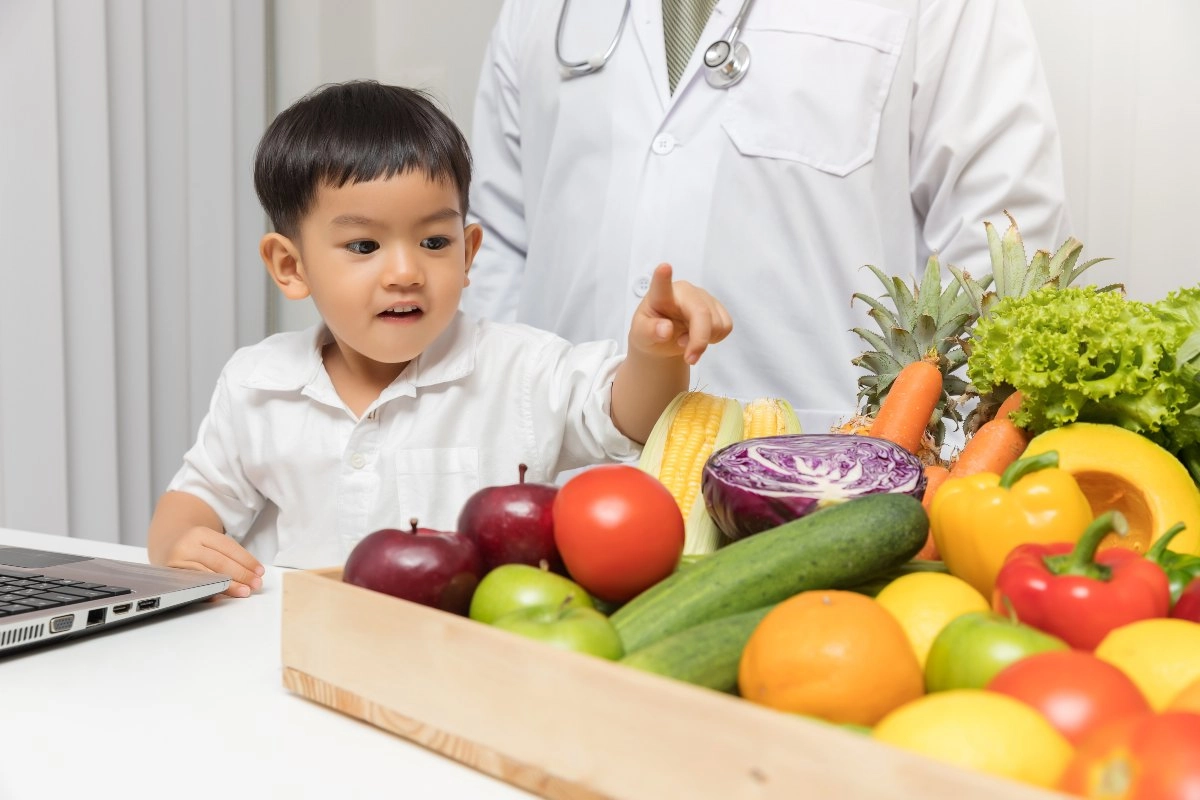 Criança apontando para uma bandeja de frutas e vegetais frescos ao lado de um médico