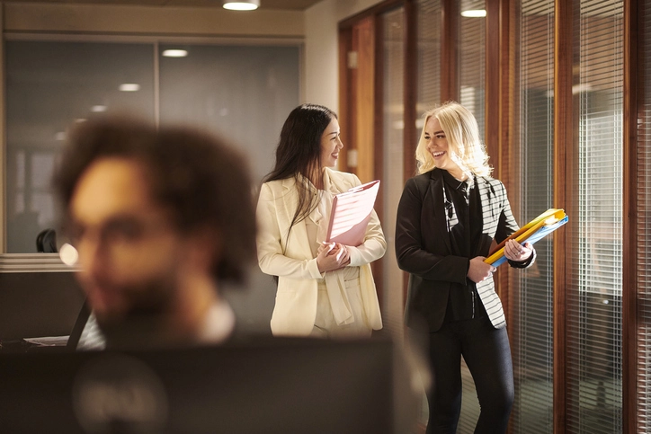Duas mulheres profissionais conversando em um ambiente corporativo segurando pastas de documentos