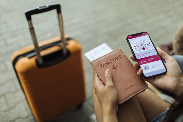 Planeje sua viagem acadêmica internacional com as dicas de internacionalização e documentos necessários para embarque Viajante segurando passaporte e cartão de embarque digital no celular com mala de viagem ao lado