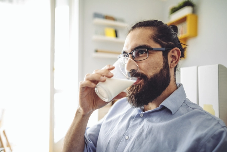 Homem com barba e óculos bebendo um copo de leite em um ambiente de escritório
