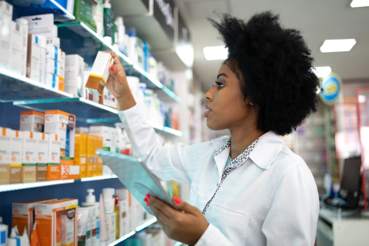 Farmacêutica negra de jaleco organizando produtos em prateleiras de uma farmácia com tablet na mão