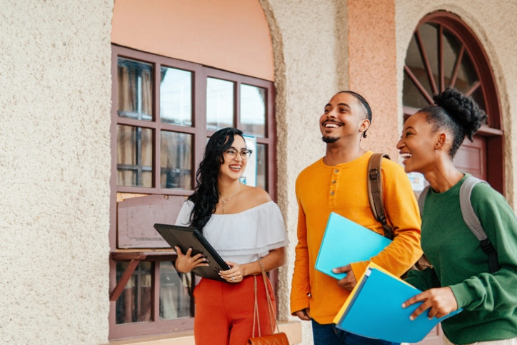 Estudantes universitários sorridentes com materiais escolares em frente a uma unidade da instituição de ensino