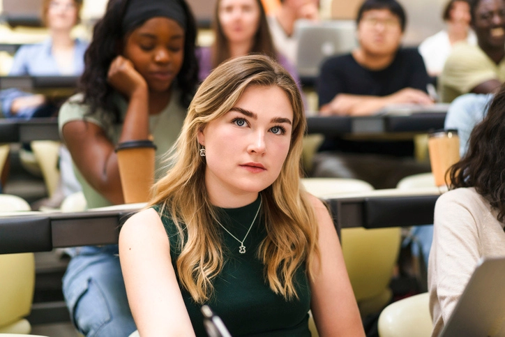 https://estaticos.animaeducacao.com.br/medias/20251008164333/tamanho_imagem_blog.webp Aluna concentrada assistindo a uma aula em um auditório universitário moderno com diversos estudantes ao redor