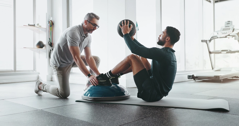 https://estaticos.animaeducacao.com.br/medias/20251010092707/GettyImages-2172581379.webp Personal trainer orientando aluno na execução correta de exercício funcional com bola bosu para fortalecimento muscular em ambiente de academia profissional