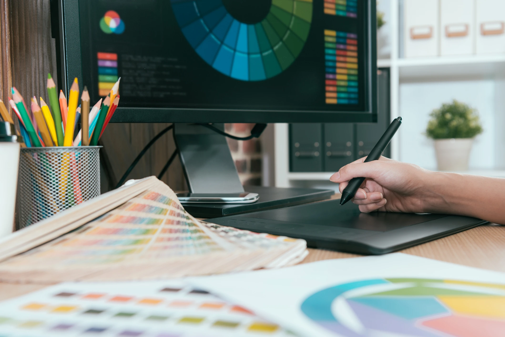 Designer gráfico trabalhando com mesa digitalizadora, círculo cromático e amostras de cores no computador Profissional de design gráfico utilizando ferramentas digitais e catálogos de cores para criar projetos visuais criativos