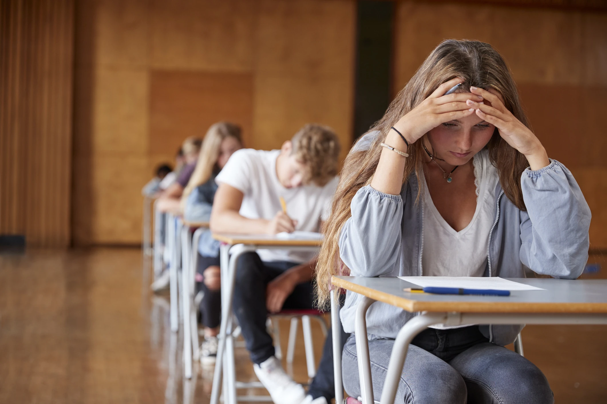 Estudante estressada fazendo prova em sala de aula com outros alunos ao fundo Estudante demonstrando estresse e ansiedade durante a realização de uma prova importante na escola ou universidade