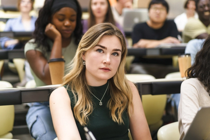 Estudante jovem loira prestando atenção em aula universitária com outros alunos ao fundo