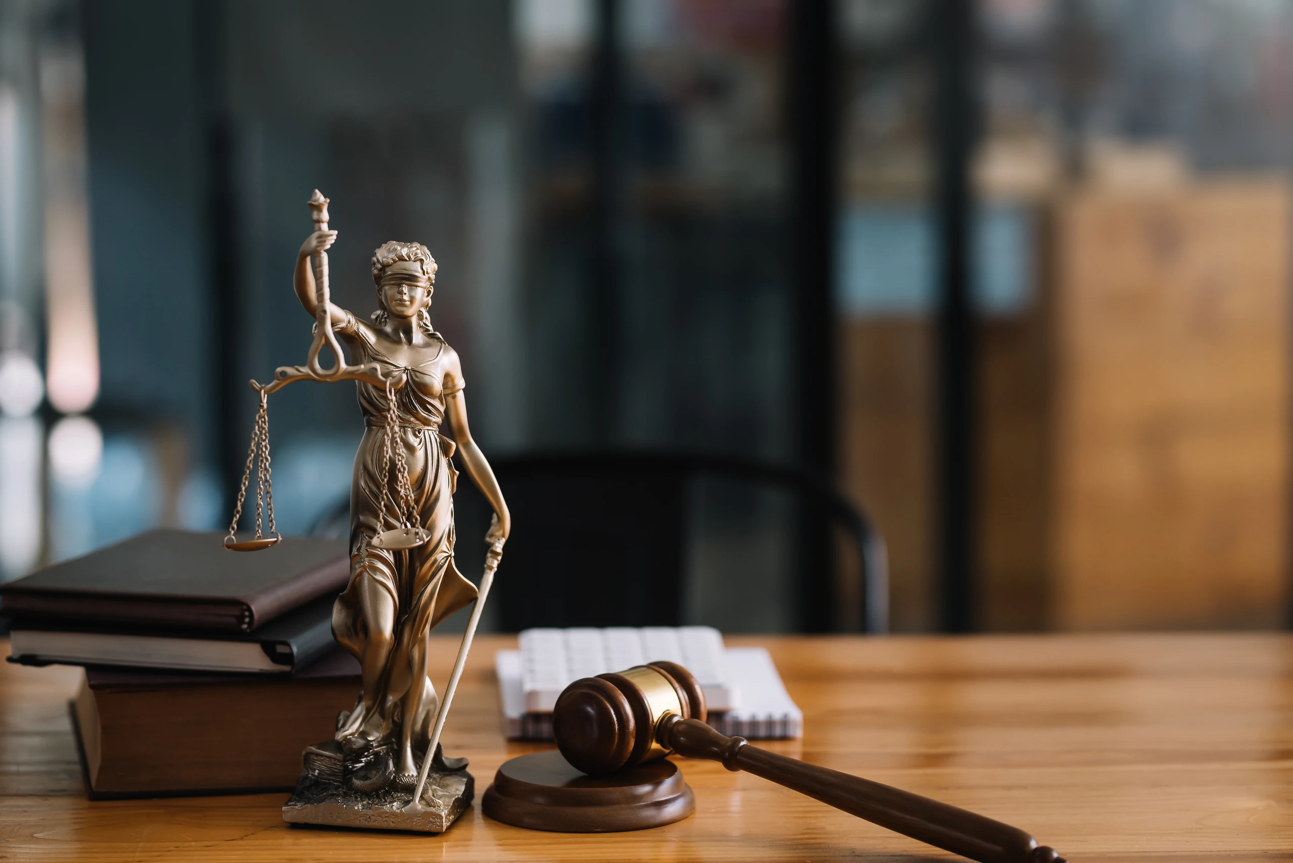 Estátua da deusa da justiça Themis com balança, livros de direito e martelo de juiz sobre uma mesa de madeira.