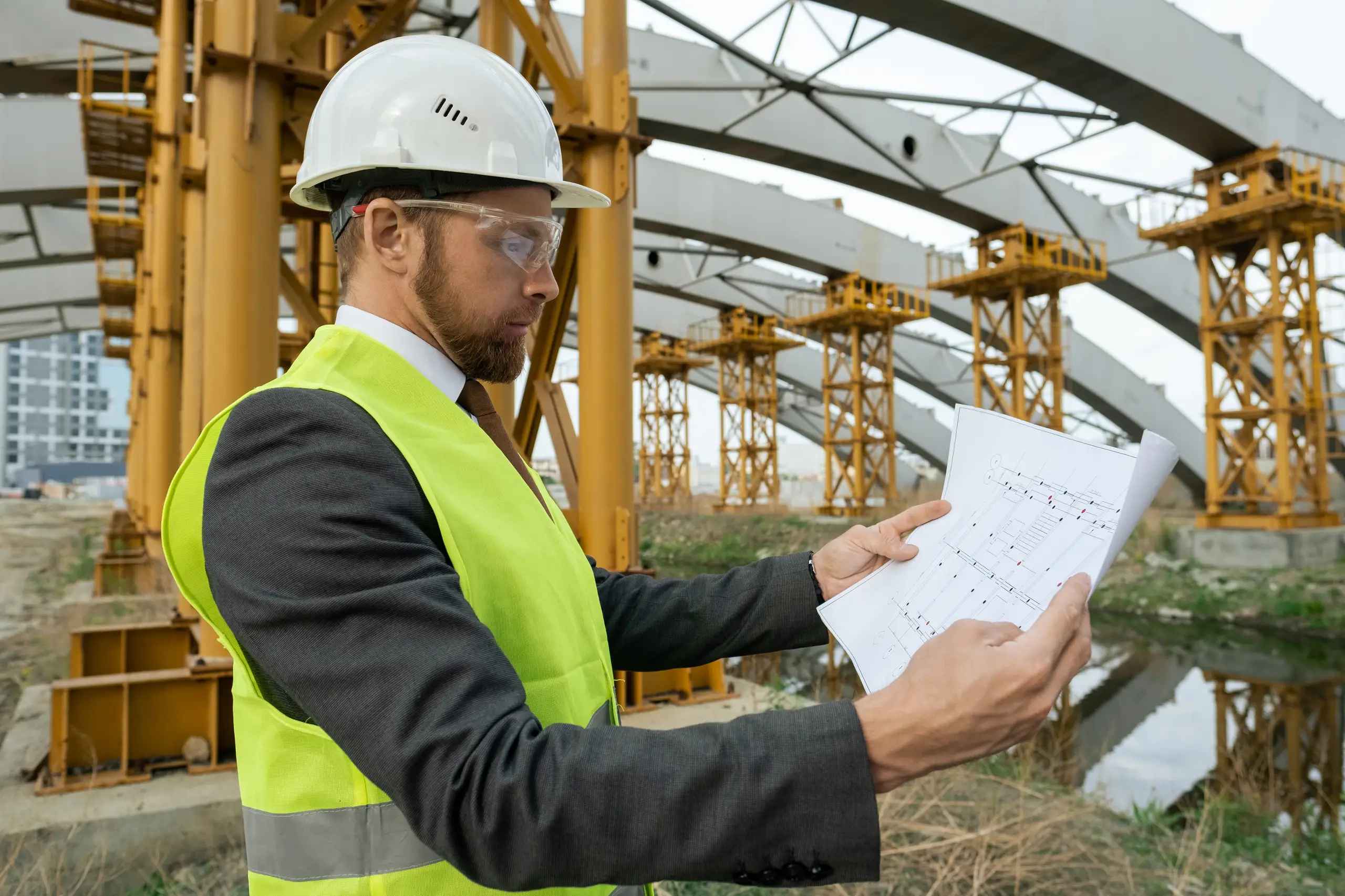 Engenheiro civil analisando planta de obra em canteiro de construção com estrutura metálica ao fundo