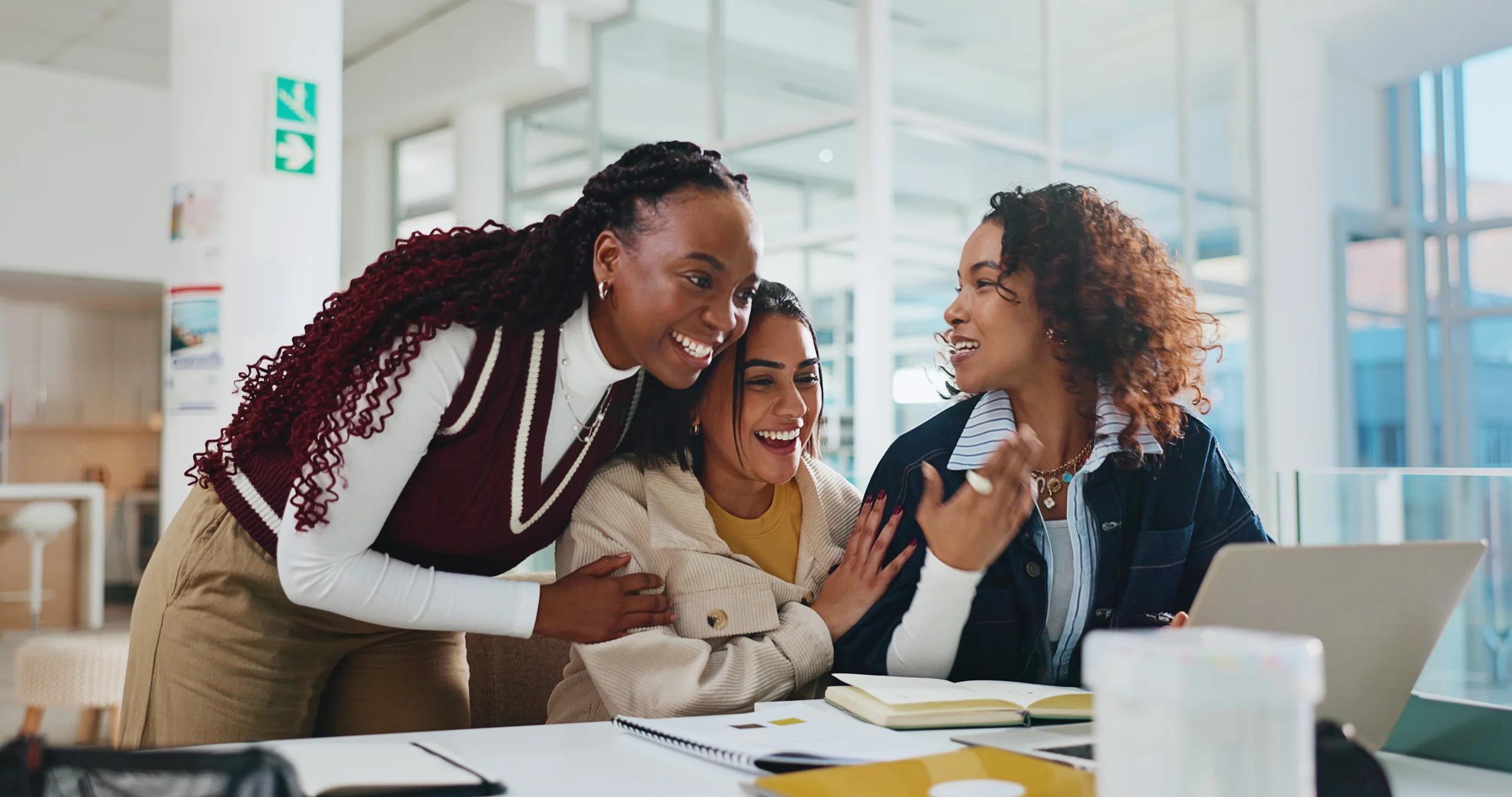 Três estudantes universitárias sorridentes colaborando em um projeto acadêmico de Letras em uma mesa de estudos moderna.