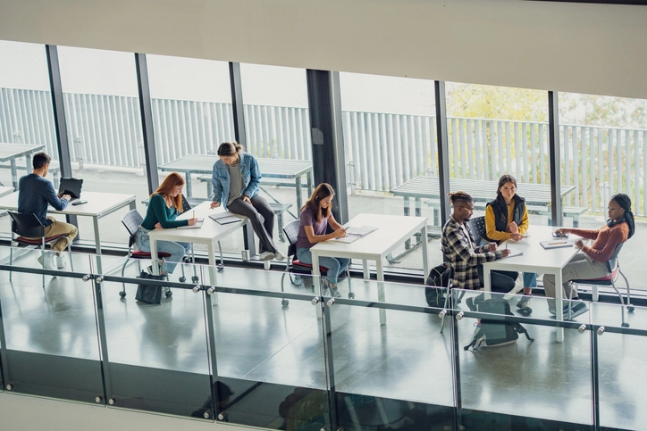 Estudantes universitários focados em seus estudos em um ambiente acadêmico moderno, colaborativo e com grandes janelas de vidro.