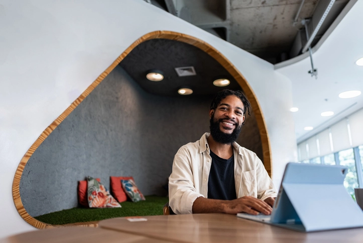 Homem negro sorridente trabalhando em um laptop em um escritório moderno com área de descompressão