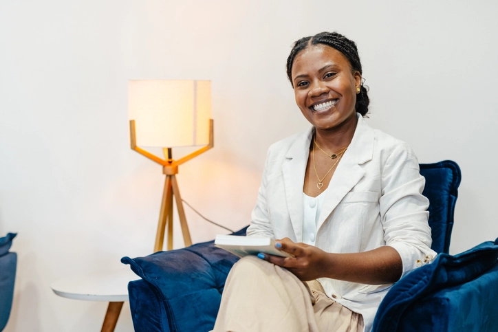 https://estaticos.animaeducacao.com.br/medias/20251216151046/GettyImages-2203722439.webp Mulher negra sorridente sentada em uma poltrona azul segurando um livro