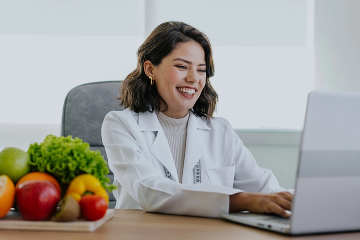 Nutricionista sorridente trabalhando em seu computador com frutas e vegetais frescos sobre a mesa
