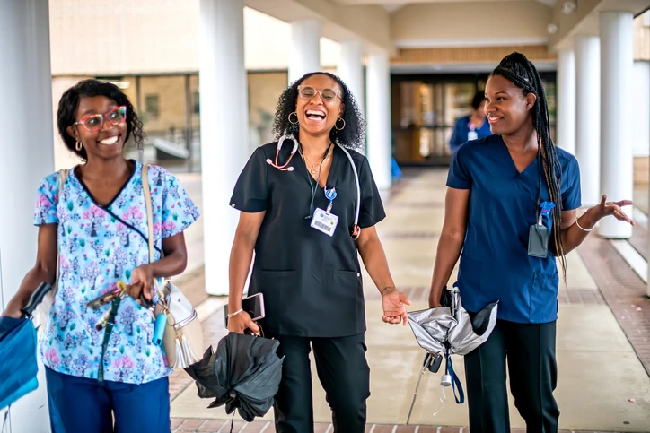 Três profissionais de saúde mulheres sorridentes caminhando juntas em um corredor hospitalar