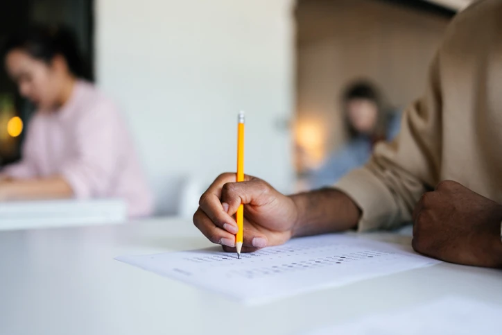 Estudante focado realizando uma prova acadêmica ou concurso público em sala de aula com outros alunos ao fundo
