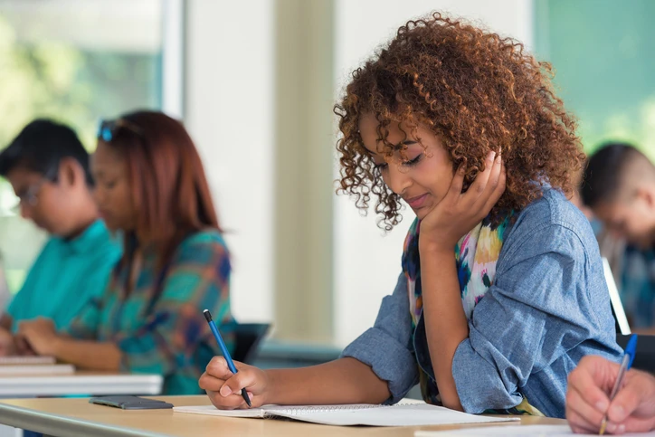 Aluna dedicada escrevendo em caderno durante aula na universidade para promover ambiente de estudo acadêmico Estudante concentrada fazendo anotações em sala de aula com outros alunos ao fundo