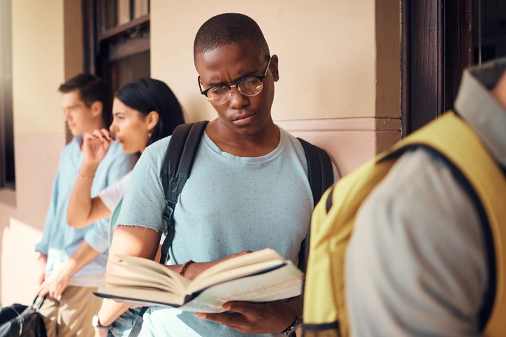 Estudante concentrado lendo livro em um campus universitário com outros alunos ao fundo