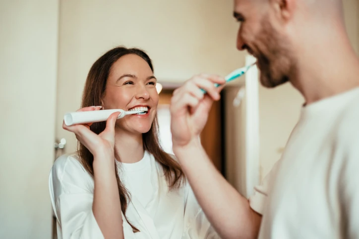 Casal sorridente escovando os dentes juntos no banheiro com escovas elétricas