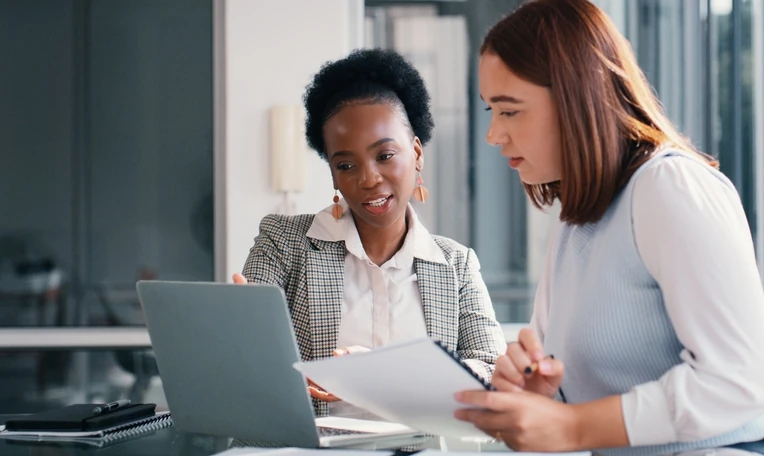 Profissionais diversas colaborando em um projeto corporativo usando notebook e documentos em um escritório moderno Duas colegas de trabalho colaborando em um projeto no escritório