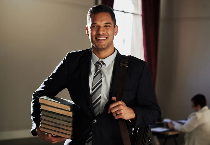 Estudante universitário sorridente vestindo terno e segurando uma pilha de livros
