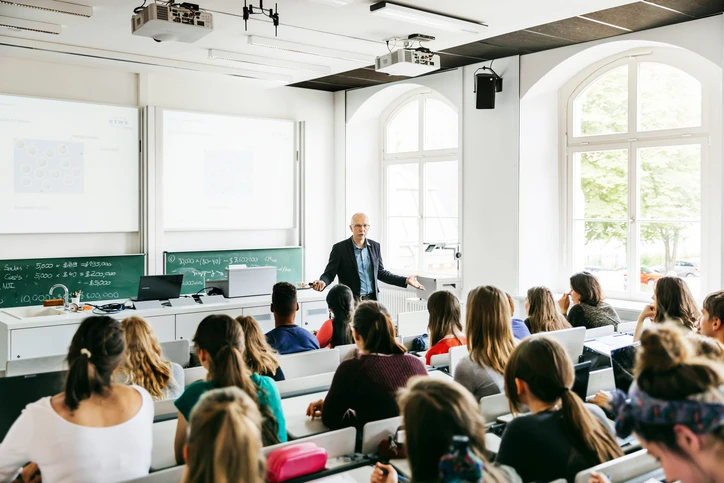 Professor universitário apresentando conteúdo acadêmico em sala de aula moderna com projetores e lousa