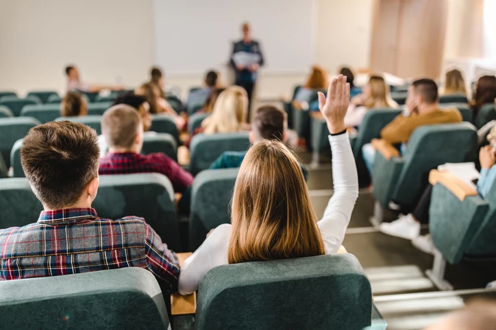 Aluna participando ativamente de uma aula universitária em um auditório com outros estudantes e um professor ao fundo