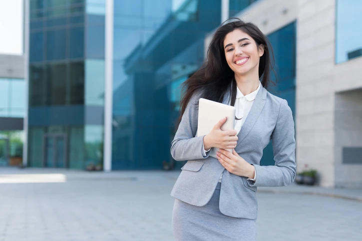 Executiva sorridente em frente a um prédio corporativo segurando um tablet