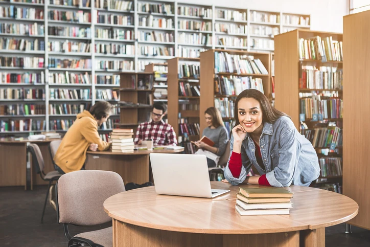 Estudante universitária sorridente estudando com laptop em uma biblioteca universitária com outros alunos ao fundo Estudante universitária sorridente usando laptop em uma biblioteca cheia de livros