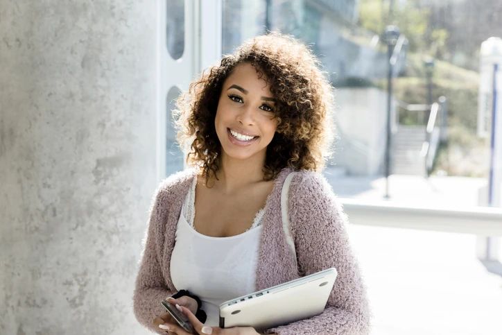 Estudante universitária sorridente segurando um notebook e um smartphone em um campus moderno