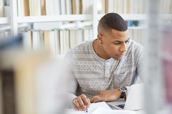 Estudante jovem focado lendo ou escrevendo em um notebook dentro de uma biblioteca
