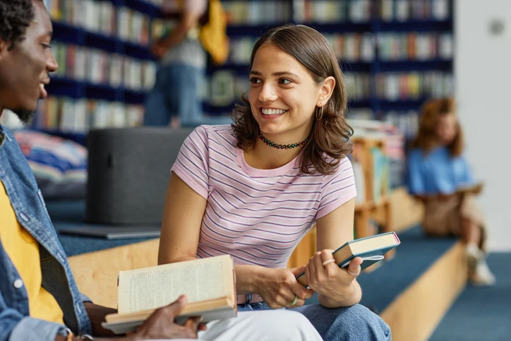 Estudantes universitários sorridentes conversando e estudando com livros em uma biblioteca acadêmica.