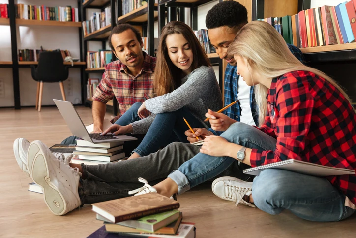 Grupo de estudantes diversos sentados no chão de uma biblioteca estudando com notebooks e livros