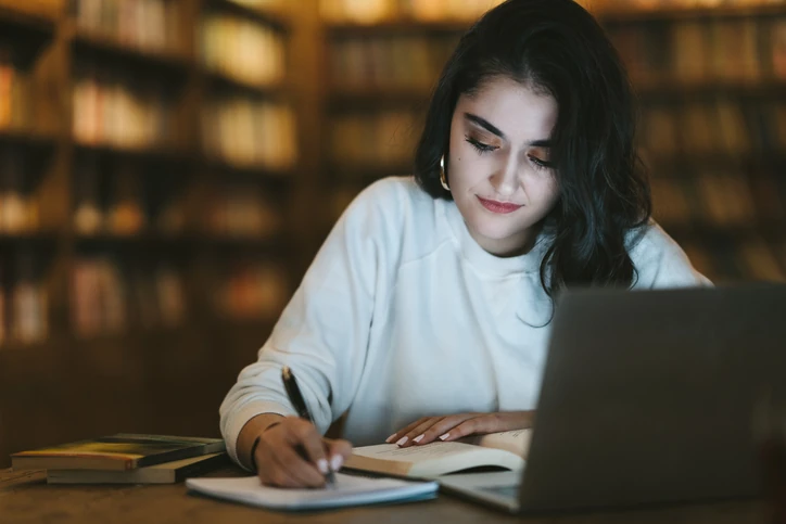 Estudante focada escrevendo em um caderno com notebook e livros em uma biblioteca