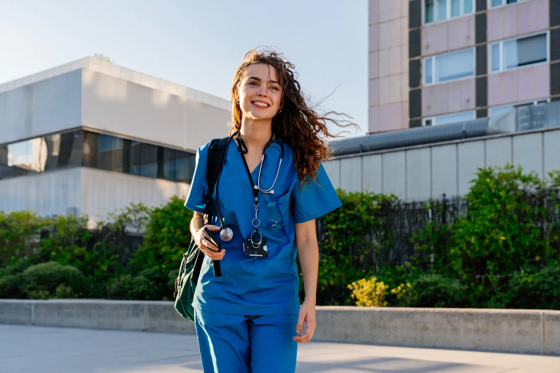 Estudante de enfermagem sorrindo vestindo uniforme azul e estetoscópio caminhando pelo campus universitário