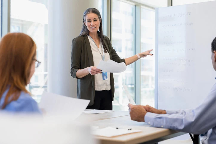Profissional da saúde apresentando casos clínicos e métricas para equipe multidisciplinar em ambiente de treinamento corporativo Professora ou supervisora apresentando dados em quadro branco para equipe em reunião